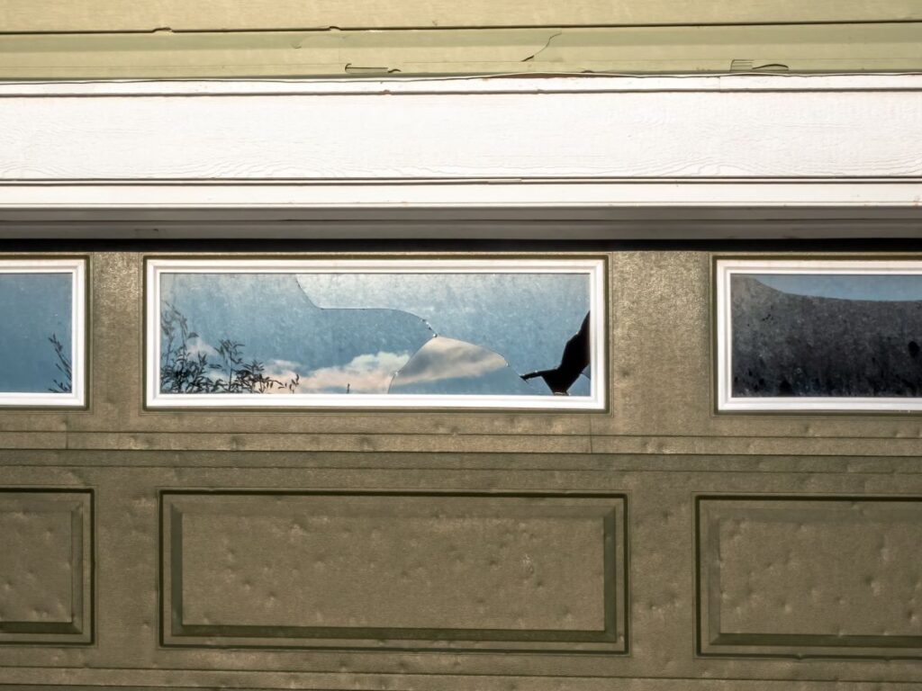 A garage door window pane is cracked with multiple impact points, indicative of hail damage
